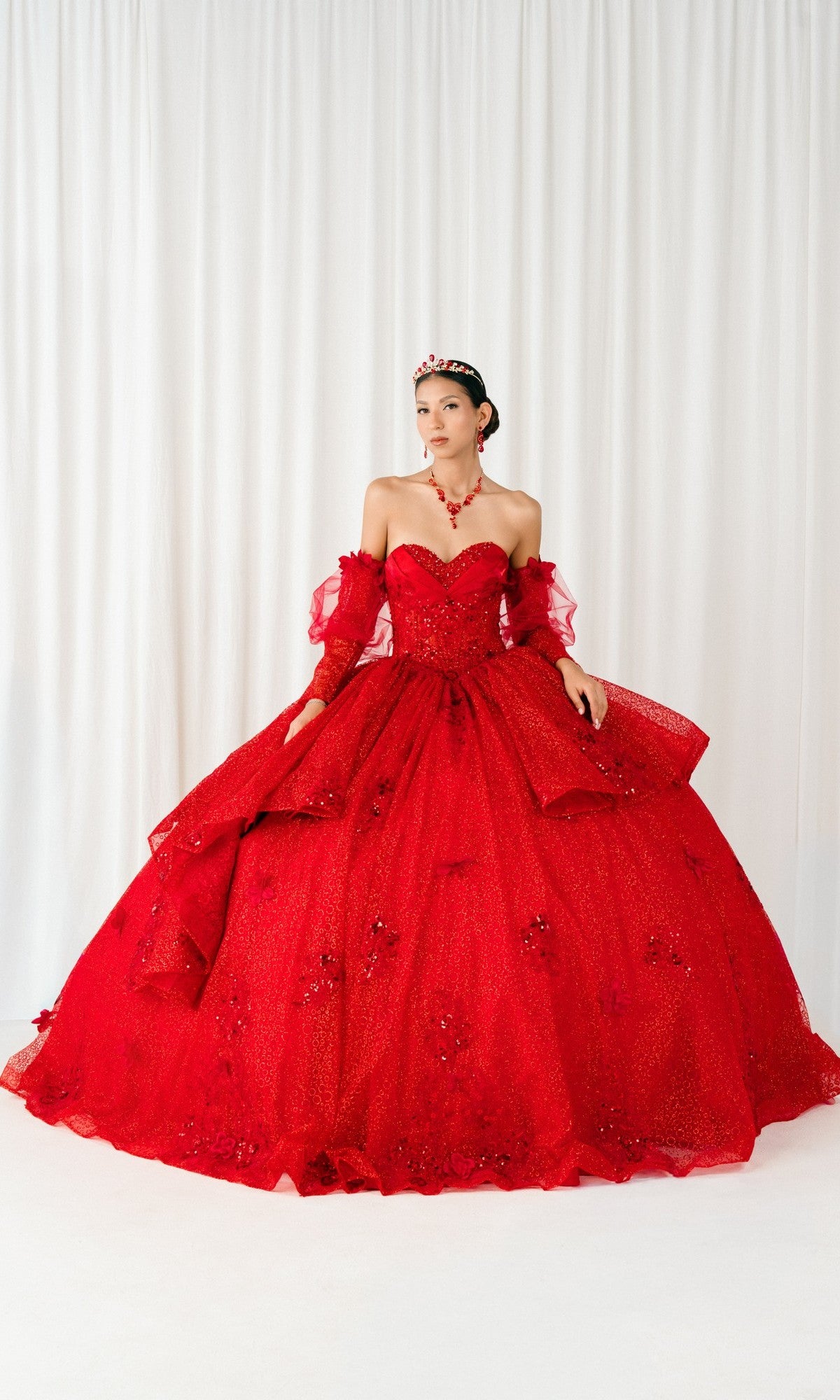 A woman wearing the red Quinceanera Dress 8385J by Cinderella Couture with a corset bodice, layered embellished skirt, matching jewelry, and a tiara stands before white curtains.
